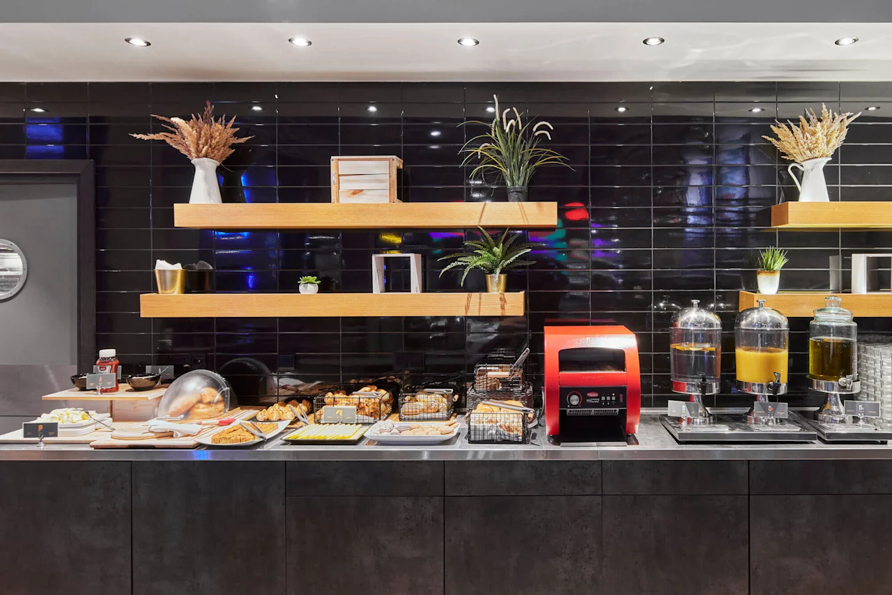 A breakfast buffet features pastries and beverages on a counter with black tiles and wooden shelves adorned with plants and decorative items.