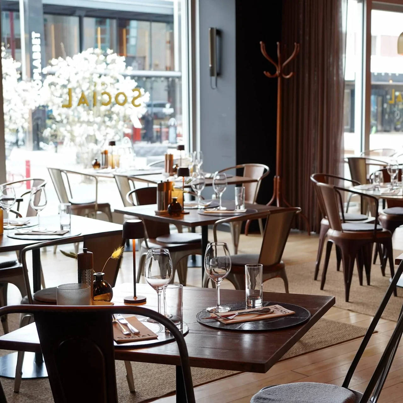 Tables are set with wine glasses and utensils in a modern restaurant, surrounded by industrial-style chairs and amber lighting. The window shows "SOCIAL" in reverse outside.