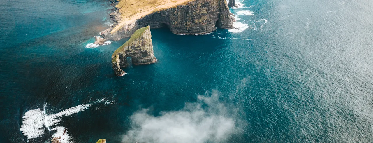 Cliffs rise steeply from the ocean, surrounded by swirling clouds and vibrant green grass. The sea crashes against the rocky shore, creating a dramatic coastal landscape.