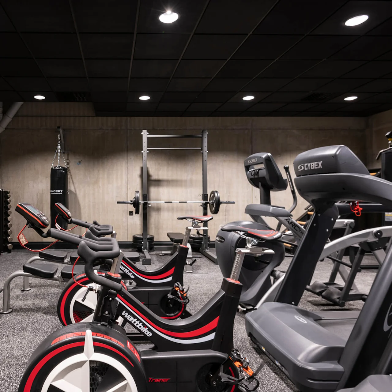 Exercise equipment stands unused in a gym. Dumbbells are on racks, stationary bikes and treadmills are nearby. The room has concrete walls and is dimly lit.