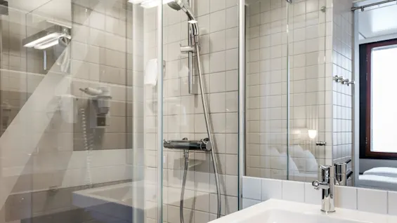 A white sink and chrome faucet with a wall-mounted shower in a tiled bathroom. Glass shower enclosure reflects the room, including a wall-mounted hairdryer.