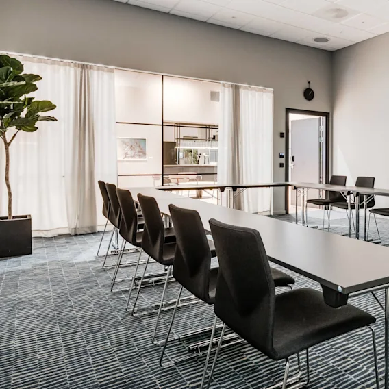 A U-shaped arrangement of black chairs surrounding tables, in a carpeted conference room with white walls and a potted plant. Beyond, glass wall reveals a kitchenette.