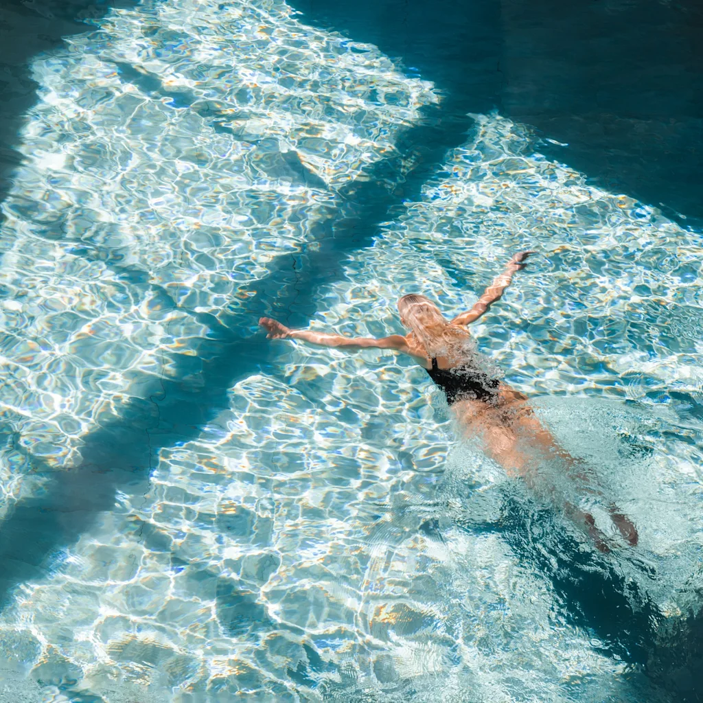 En person svømmer afslappet i en soloplyst pool og skaber krusninger. Vandet reflekterer skinnende lysmønstre, der understreger en fredelig atmosfære.