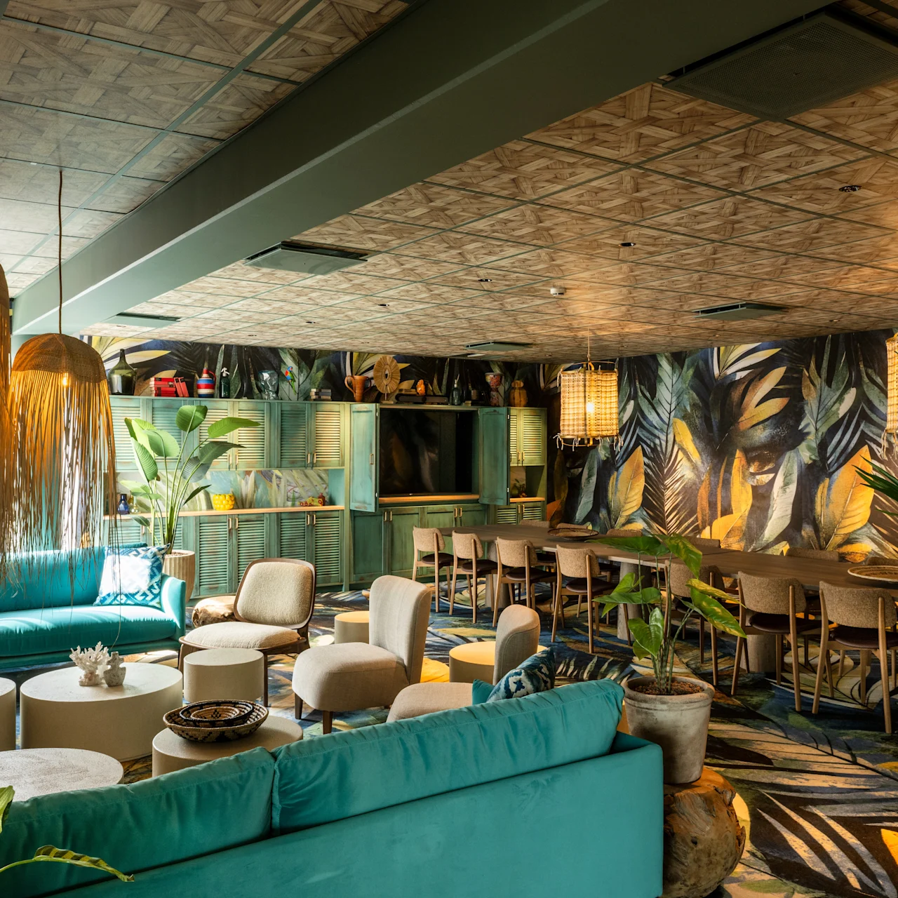 A teal sofa and chairs surround a low table, set in a vibrant room with tropical wallpaper and plants. Overhead, woven pendants cast light, creating a cozy, inviting atmosphere.