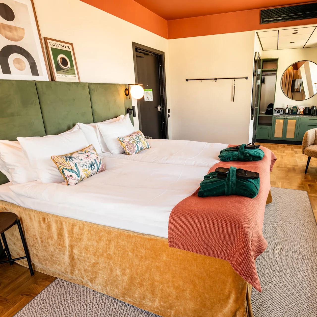 A neatly made bed with green pillows and blankets lies under framed artwork. Two green robes rest on an orange throw. A round mirror and chair are in the modern hotel room. Text: “Espresso.”