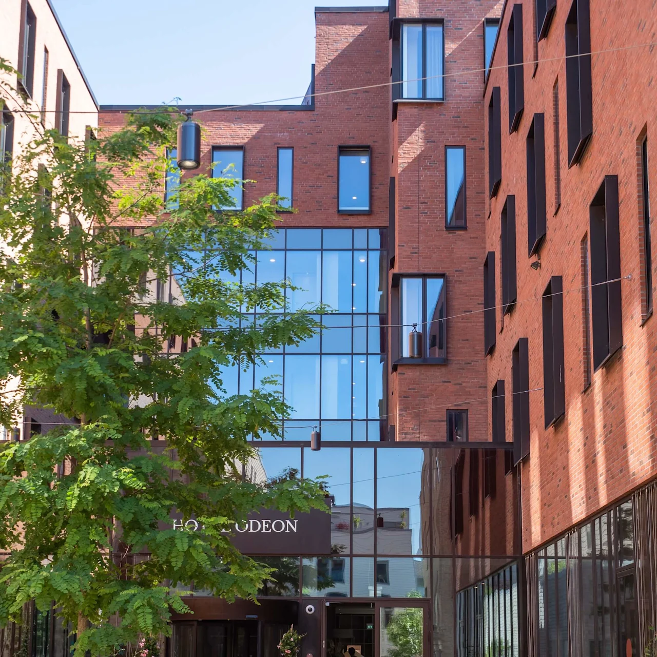 Moderni tiilirakennus, jossa on lasinen sisäänkäynti, sanoo yläpuolella "HOTEL ODEON". Puu ja pyöreä istutus koristavat tiilikatriikkia. Penkit reunustavat kävelytietä.