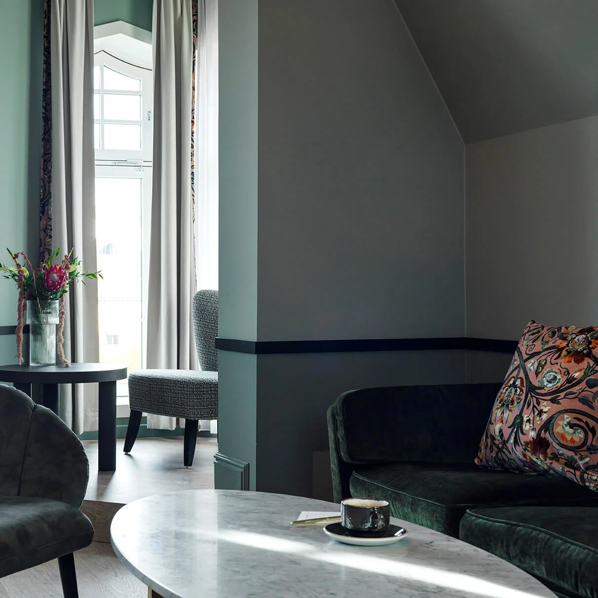 A velvet chair rests beside a sleek marble table holding a coffee cup in a softly lit room with large windows draped in curtains. A flower vase sits on a side table.