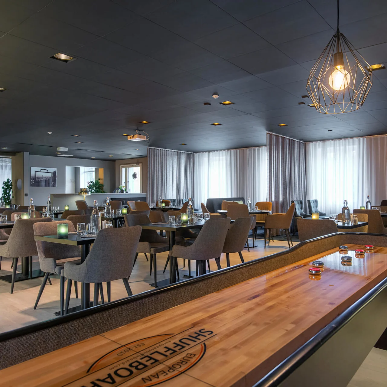 Shuffleboard på bord, ospelat, i modern restaurang med eleganta stolar och bord dukade med vattenflaskor och glas, hängande lampor ovanför, och stora fönster med skira gardiner.