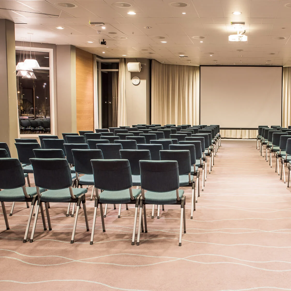 Rader med tomme stoler vender mot en stor projektorskjerm i et godt opplyst konferanserom med nøytrale vegger og gardiner.