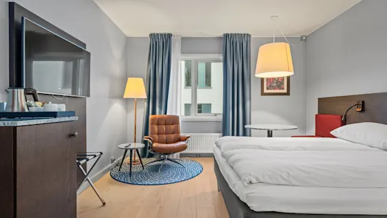 Et ryddeligt hotelværelse med seng, trægulv, lænestol og bord under en lys loftslampe. Blå gardiner dækker vinduet, og et TV er monteret på væggen.