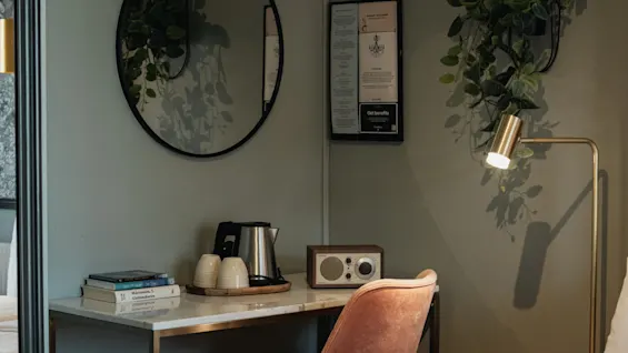 Et hyggeligt hotelværelseshjørne med skrivebord, stol og seng. En elkedel og radio står på skrivebordet, og planter pryder væggene. Få fordele.