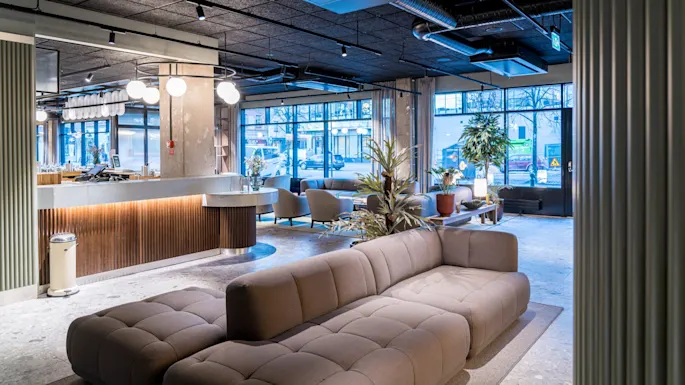 En moderne lounge har en stor, polstret sofa i et romslig, godt opplyst rom med store vinduer, omgitt av potteplanter og stilige lysarmaturer.