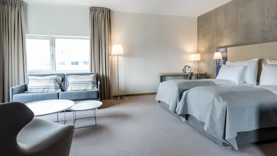 A hotel room features two neatly made twin beds, a gray sofa with striped pillows, and a white coffee table. Floor lamps and a desk with a coffeemaker complete the modern decor.
