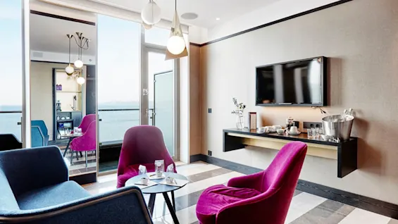 En moderne stue med en lilla stol og blå sofa vendt mod et monteret TV. En glasdør åbner ud til en balkon med udsigt over vandet. Et sidebord har to glas.