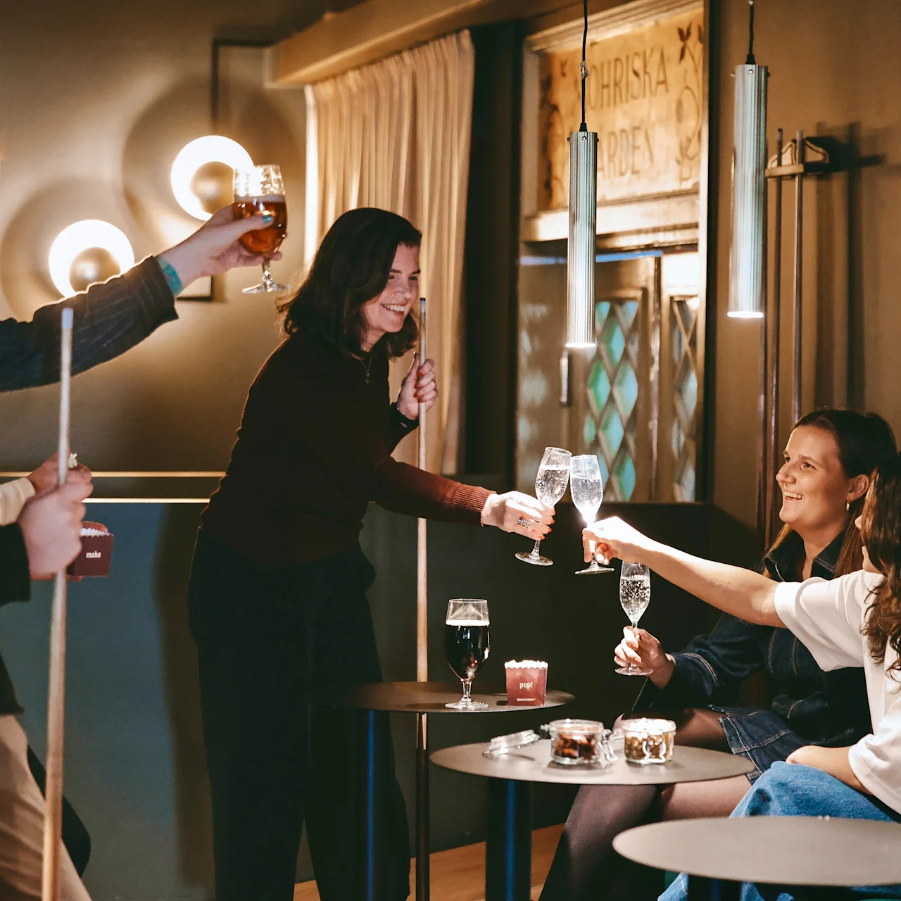 Five people toast with drinks at small round tables, creating a lively atmosphere in a warmly lit bar. Wall lights and a pool cue add ambiance.