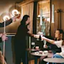Five people toast with drinks at small round tables, creating a lively atmosphere in a warmly lit bar. Wall lights and a pool cue add ambiance.