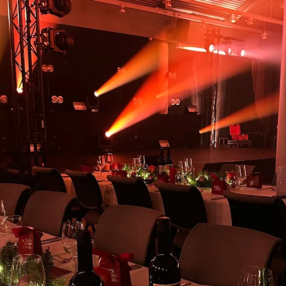 En festsal pyntet til julebord med dekkede bord, røde lys og varm oransje scenebelysning i bakgrunnen.