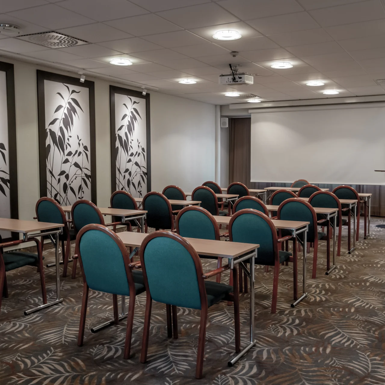 Rows of green chairs face tables in an empty conference room with leaf-patterned panels on the walls and a projector screen at the front of the room.