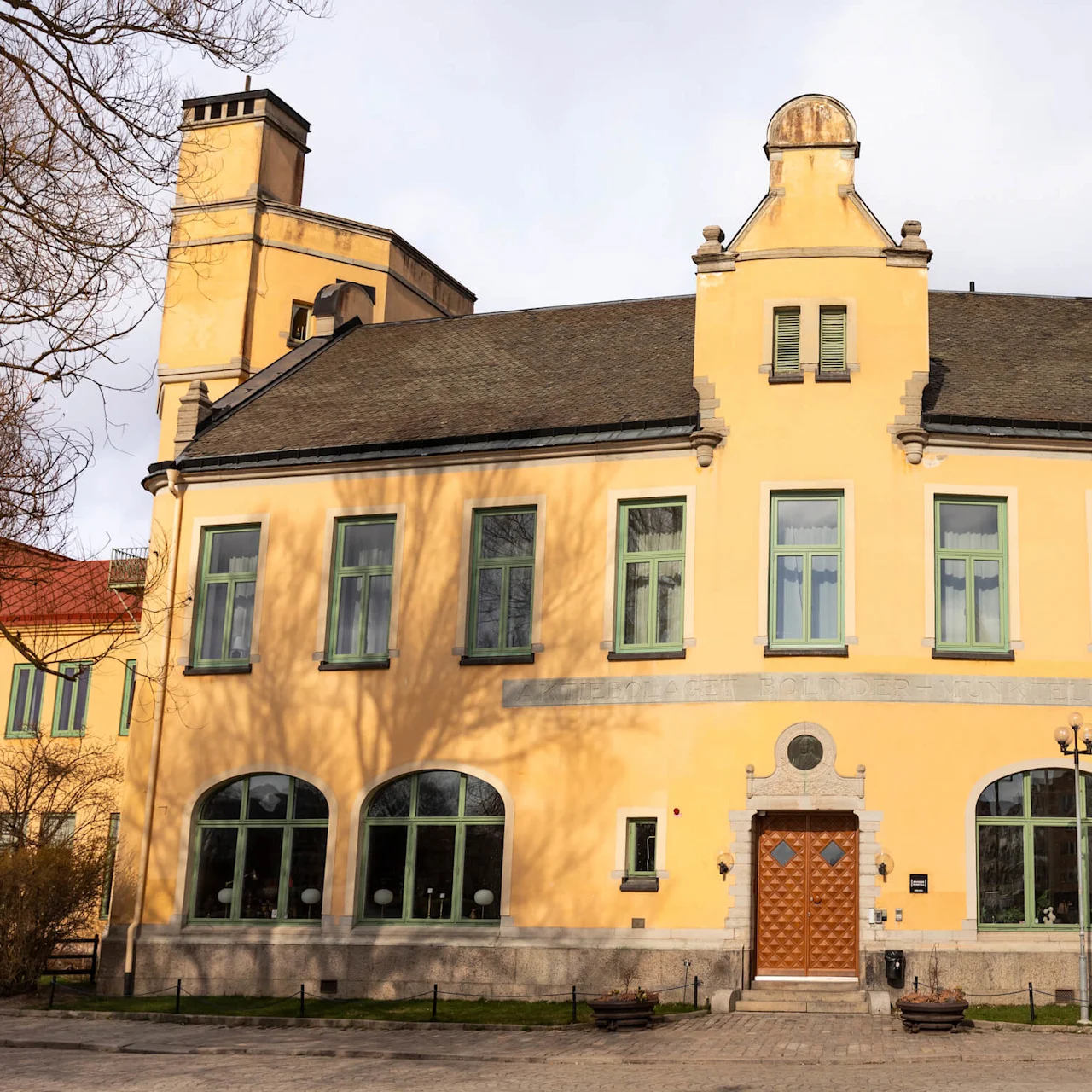 En gul byggnad med grönkantade fönster och en mönstrad trädörr står på en kullerstensgård. Ovanför ingången står det otydligt "ASCHEBERGAGYMNASIET BOLIDENHUSE." En klocka syns till höger.