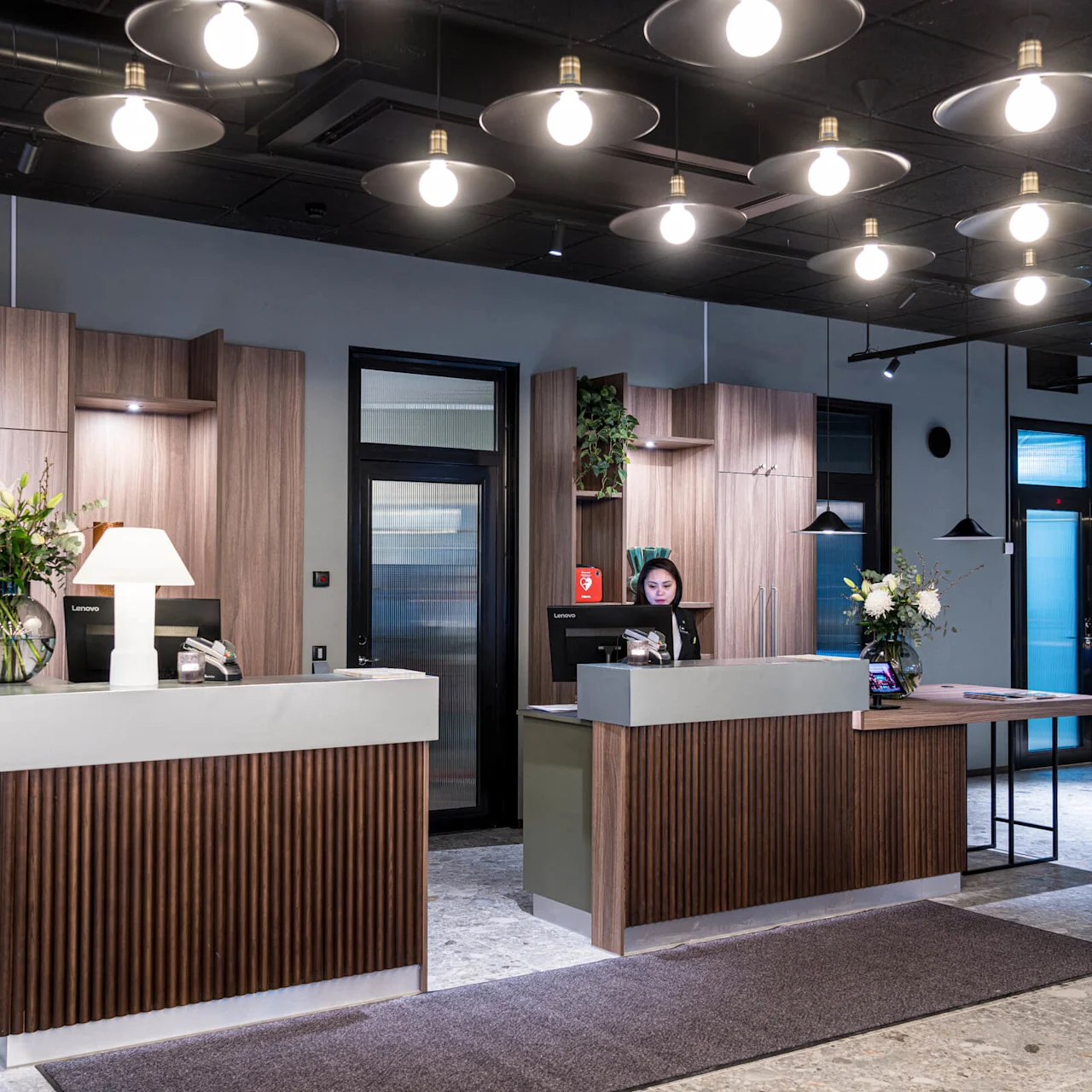 Reception desks with wooden panels stand in a modern lobby. A person at one desk works on a computer. Overhead lights illuminate the space, decorated with flowers and a lamp.