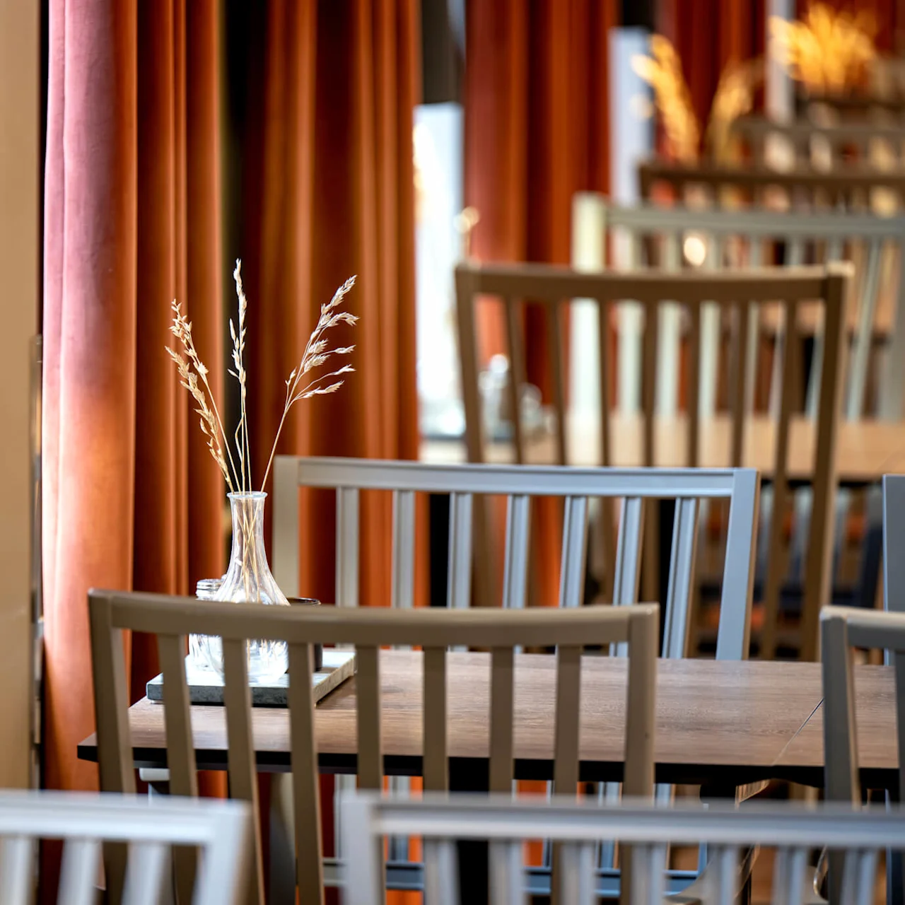 Wooden chairs and tables in a warmly lit restaurant setting; a small vase with dried plants on one table.