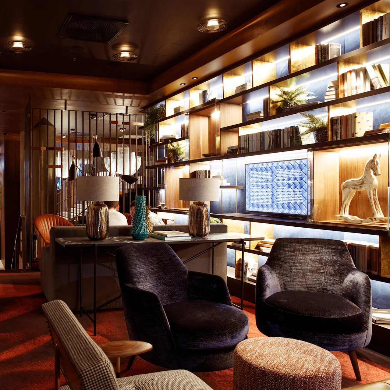Luxurious lounge featuring plush chairs and dim lighting, surrounded by shelves with books and decorations, creating an inviting atmosphere. Plants and artworks enhance the sophisticated room design.