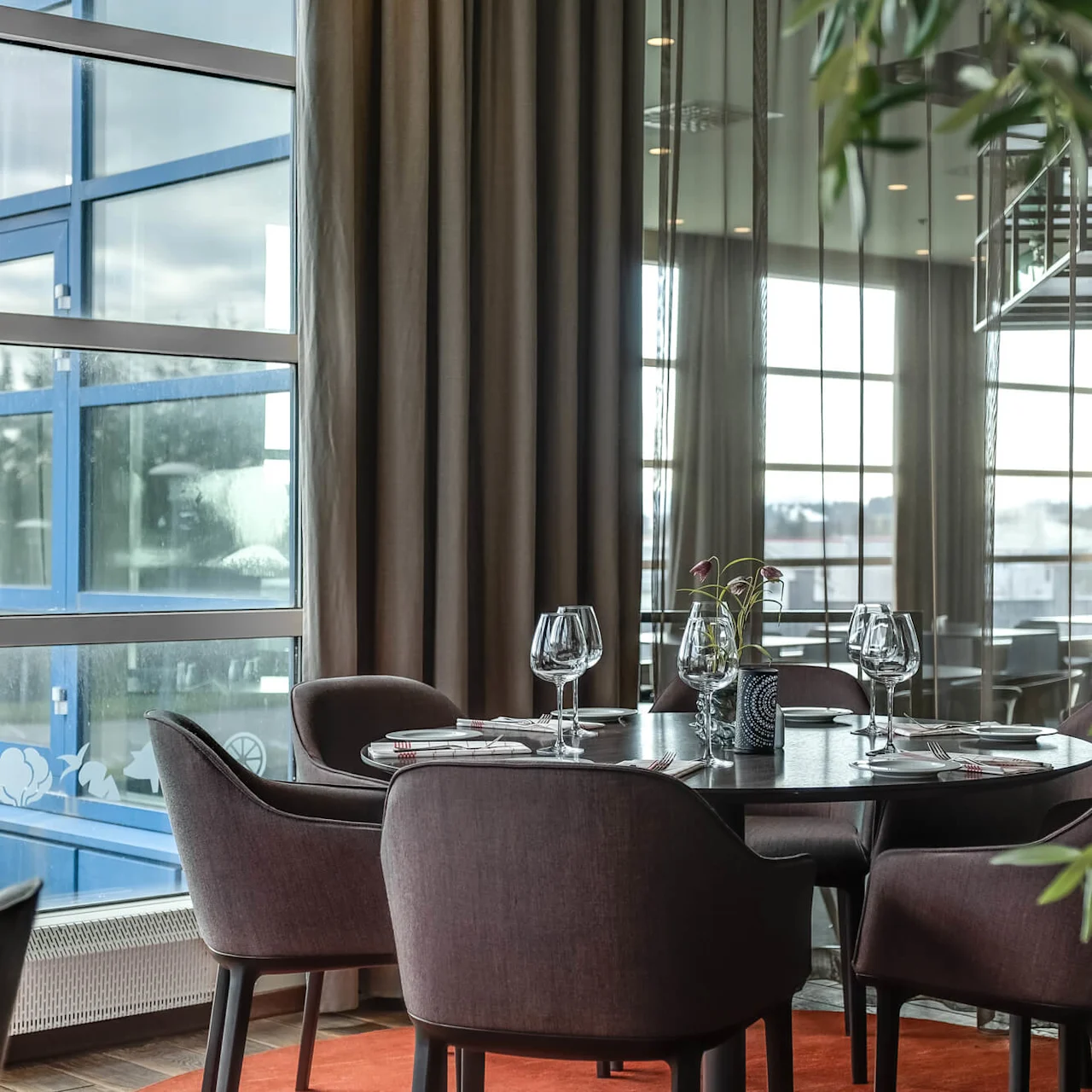 A round dining table set for four, with empty wine glasses and cutlery, in an elegant room with large windows and curtains.