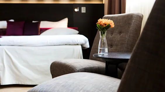 Viihtyisä hotellihuoneen oleskelualue, jossa on ruskeat nojatuolit, pieni pöytä kukkineen ja sänky taustalla.