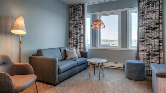 Gray sofa sits under a bright lamp, accompanied by a round table and armchair, in a softly lit room with large windows and patterned curtains.
