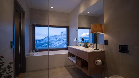 A luxurious bathroom features a bathtub and modern vanity with a lamp, set against a large window displaying a snowy mountain landscape.