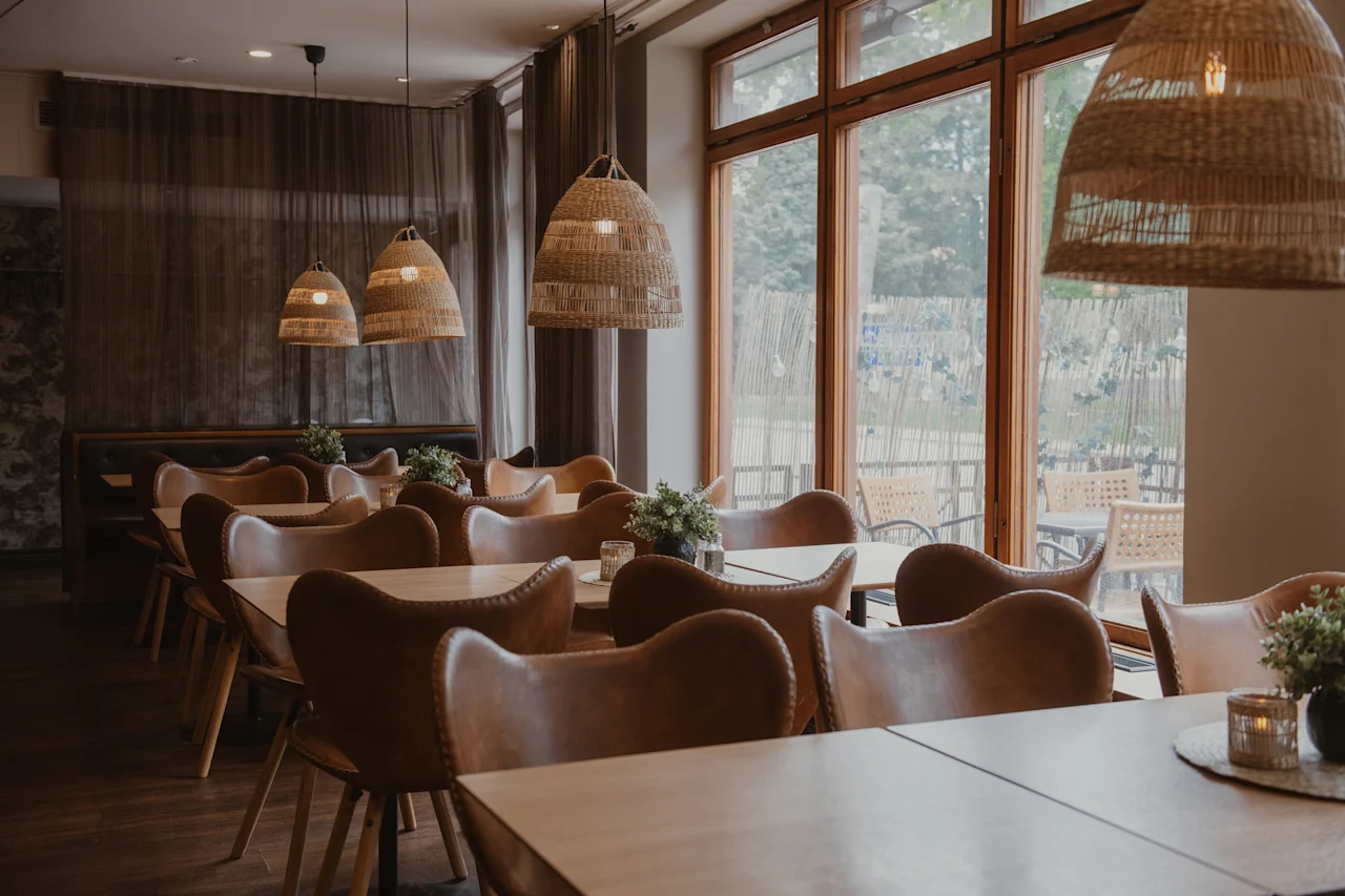 A cozy restaurant dining area with wooden tables, leather chairs, and woven pendant lights. Large windows overlook an outdoor space.