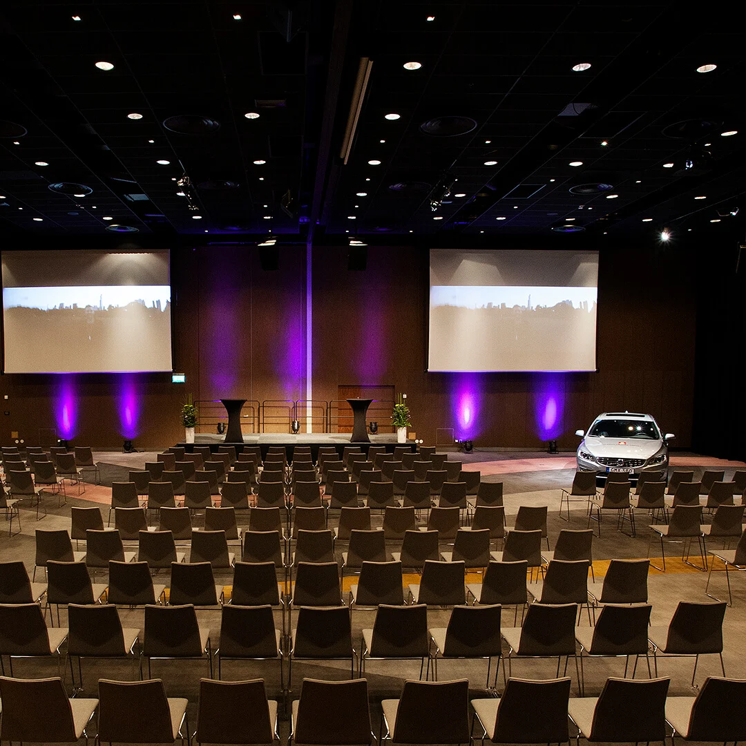 A large conference room with rows of empty chairs facing a stage. A car is parked near the stage under purple lights, with projections on the wall.
