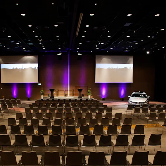En stor konferenssal med rader av tomma stolar som vetter mot en scen. En bil står parkerad nära scenen under lila ljus, med projektioner på väggen.