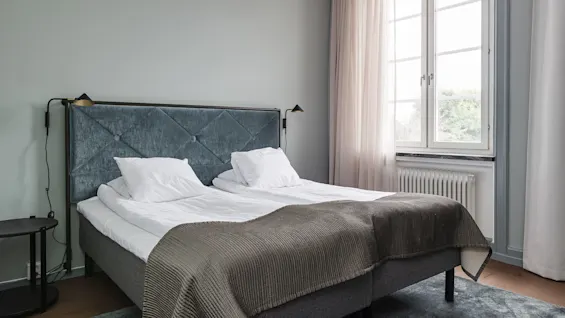 A neatly made bed with a dark grey headboard, in a room with soft light from a window on a sunny day.