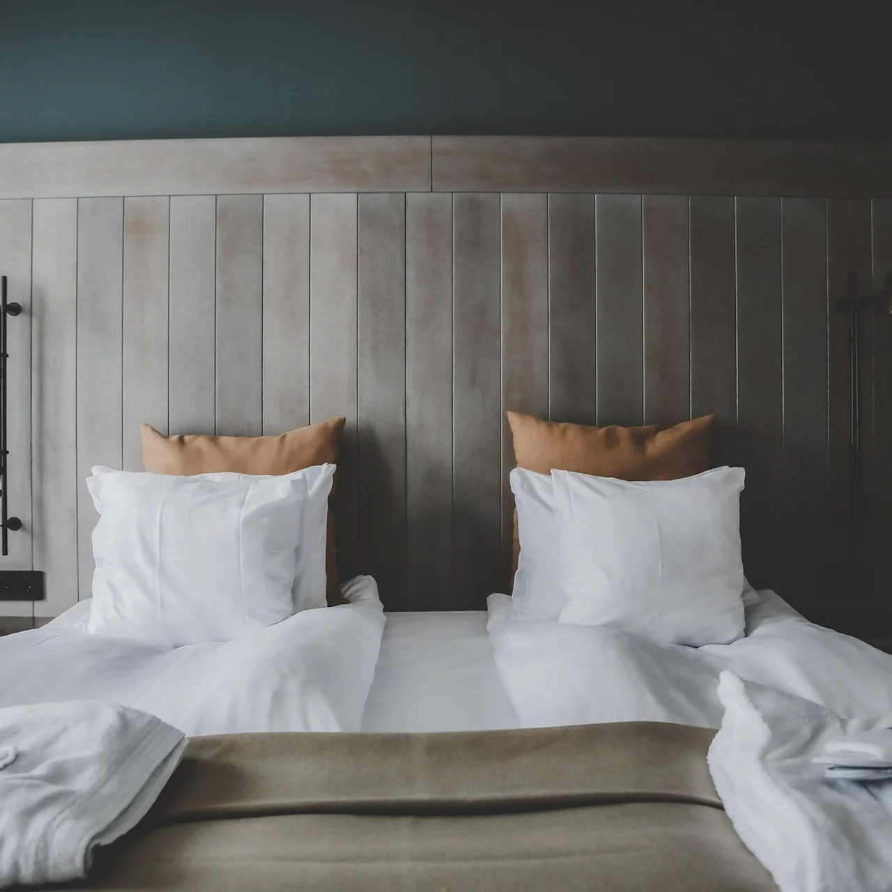 A neatly made bed with white linens and beige pillows sits beneath a wooden headboard, flanked by two wall-mounted copper lamps, creating a cozy, minimalist room setting.