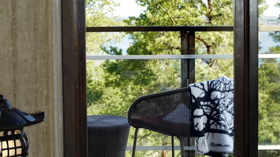 En stol med et dekorativt pledd har utsikt over en balkong og grønne trær fra innsiden av et moderne rom.