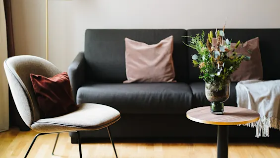 En grå sofa med beige puder står i en hyggelig stue, ledsaget af en moderne stol, et rundt træbord med en vase med grønt, og en stålampe.