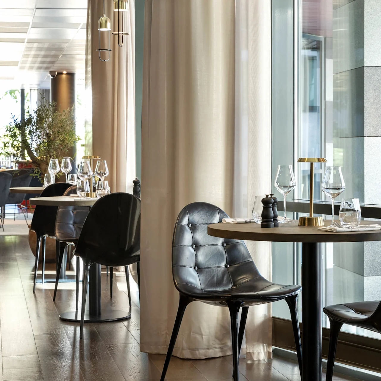 Tables are elegantly set with wine glasses and black chairs, located by large windows in a modern, well-lit restaurant with soft curtains.