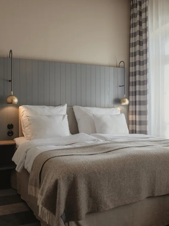 Et koselig hotellrom med en stor seng, hvite puter og et varmt teppe. Lys strømmer inn fra et vindu med rutete gardiner.
