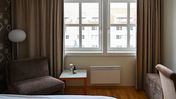 Ett mysigt hotellrum har en säng, fåtöljer och ett litet bord med en vinflaska och glas. Ljusa fönster och tunga gardiner släpper in mjukt dagsljus.