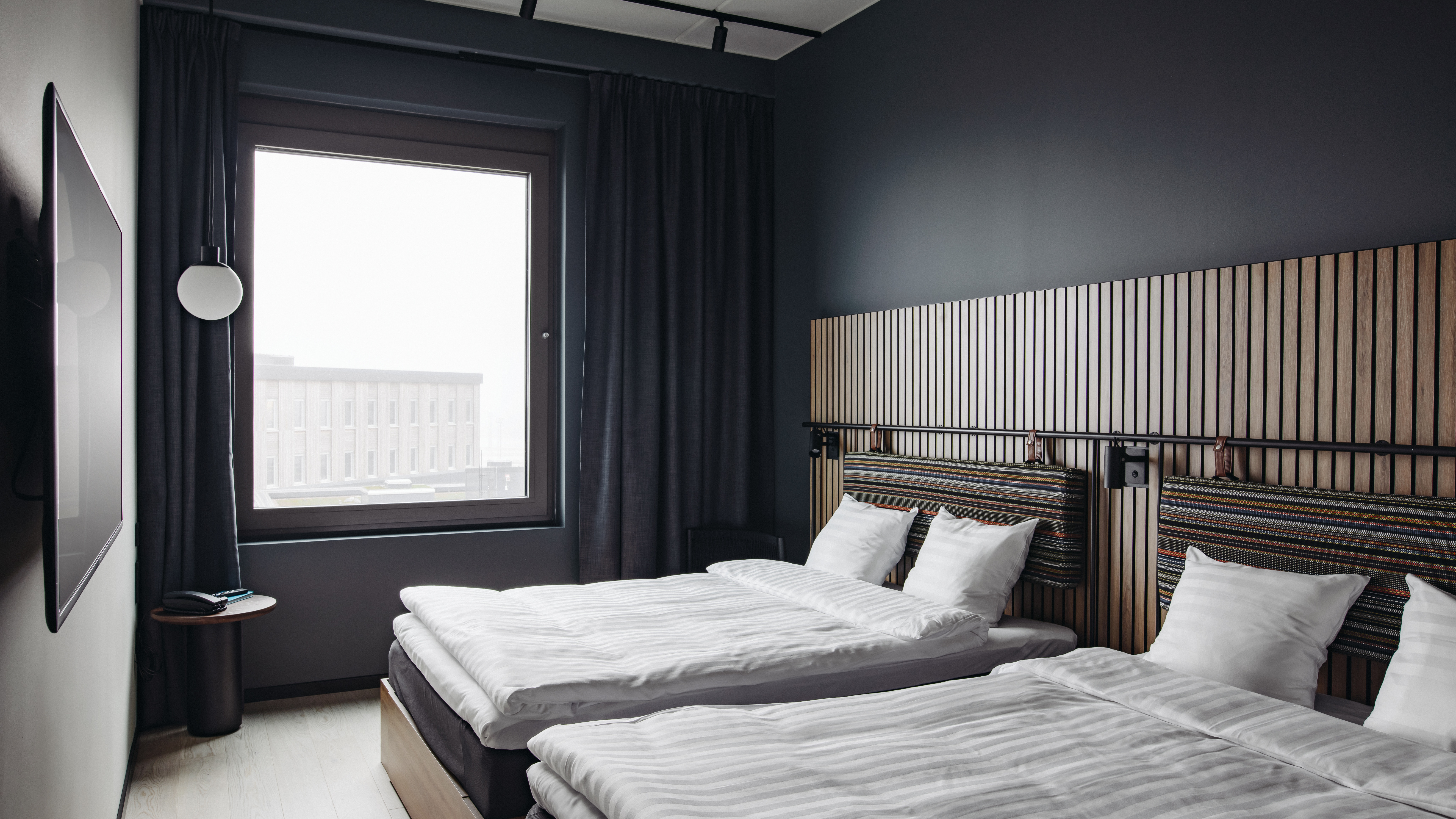 Et moderne hotelværelse med to senge, mørke vægge og et stort vindue. Værelset har en sengegavl i træ og et vægmonteret TV.