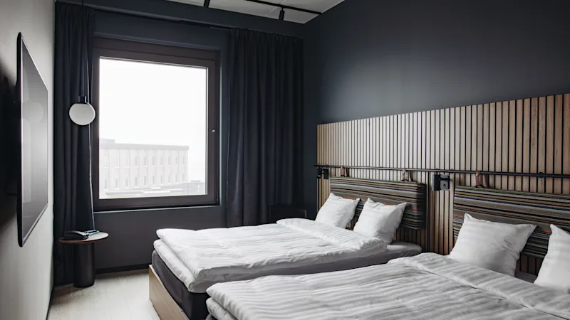 Et moderne hotelværelse med to senge, mørke vægge og et stort vindue. Værelset har en sengegavl i træ og et vægmonteret TV.