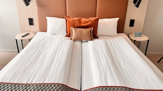 A neatly made bed with white sheets and brown-orange pillows sits between two bedside tables holding lamps and a remote control; a no smoking sign is visible.