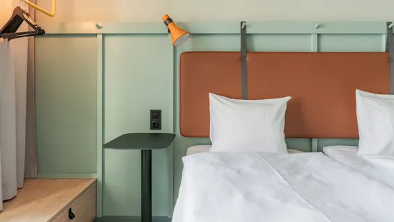 En sirligt redt seng med hvidt sengetøj er placeret mod en lysegrøn væg med en orange sengegavl. I nærheden ses bøjler og en tændt lampe.