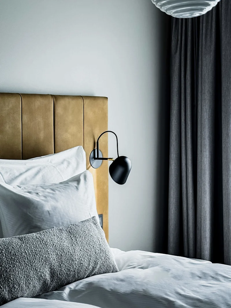 En säng med vita lakan och en grå texturerad kudde står mot en solbränd klädd huvudgavel. En svart vägglampa är fäst bredvid den, under en rund taklampa.