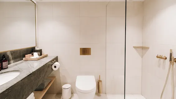 Ett modernt, rent badrum med en marmorbänk, vit toalett och glasdusch med guldfärgade armaturer.