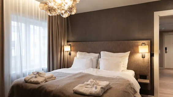 A cozy hotel room at Home Hotel Grand Bodø with a large bed, soft linens, and bathrobes, illuminated by a stylish chandelier.
