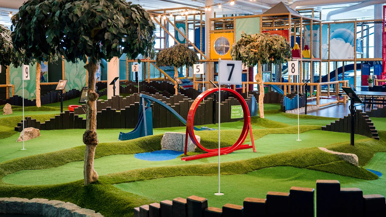 Inomhus minigolfbana har numrerade hål, konstgräs och livfulla hinder som ramper och loopar. Dekorativa träd och en rymlig lekstruktur i bakgrunden fullbordar den rekreativa miljön.