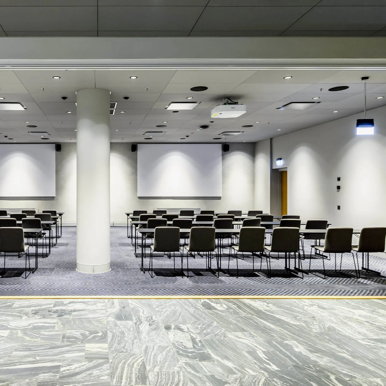 Rows of empty chairs face two blank projection screens in a spacious conference room with overhead lighting and a central support pillar.
