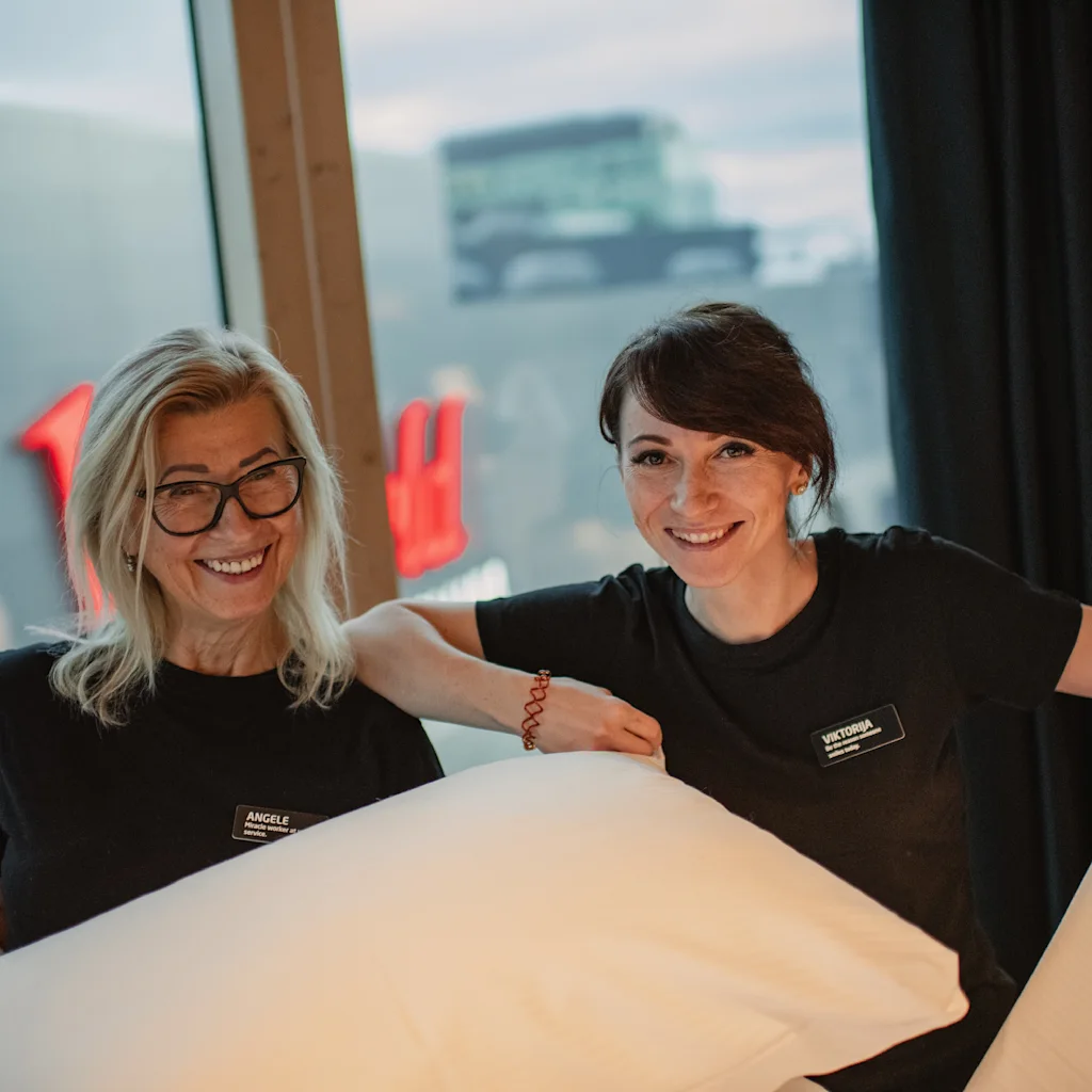 Two smiling women hold pillows in a warmly lit room, standing near a window. They wear black shirts with name tags, suggesting they're preparing a space, possibly in a hotel.
